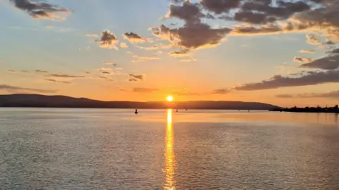 Billy Darroch A calm sunset over a wide body of water, with the sun low on the horizon casting a golden reflection across the surface, silhouetted hills in the distance, and scattered clouds lit by warm orange and pink tones.