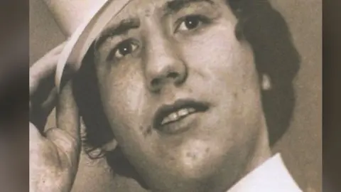 Columba McVeigh, looking to the right of camera, with his mouth slightly open. He is wearing a white shirt and his hand is holding onto a had which is on his head.