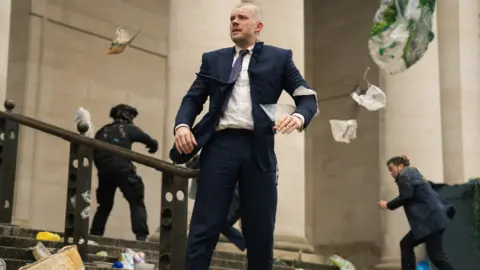 BBC Studios/Bad Wolf/James Pardon Tovey no personagem Barclay parado na escadaria do lado de fora do Museu Nacional de Cardiff. Ele veste um terno azul-marinho e parece angustiado enquanto o lixo circula pelo vento. Atrás dele, um policial armado pode ser visto e um homem de terno correndo em perigo. 