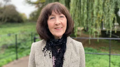 Jackie Brockway who has dark brown bobbed hair and is wearing a tweed jacket and black scarf is standing in front of a willow tree on a lawn.