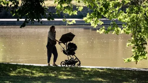 PA Media A woman pushing a buggy along a path beside a pond in a park. She and the buggy are both silhouetted by the sun. It's a bright day, with the sun shining off the water and on the leaves of trees.