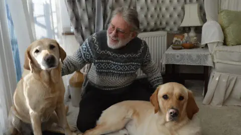 Contributed A man with grey hair kneels on the floor of a living room with two golden Labradors next to him. The one on the left is sitting on its back paws while the other is completely lying down. The man is stroking each dog and smiling at the one on the left. 