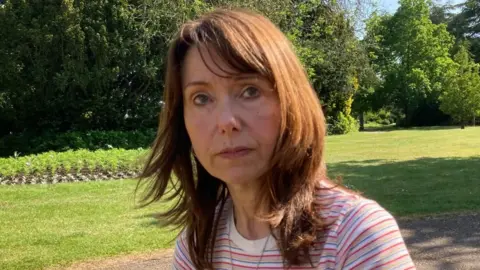 Martin Heath/BBC Angie Mower with long brown hair wearing a pink striped top sitting in a park