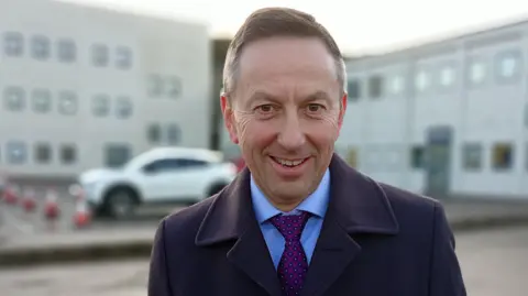 Brian Donaldson has short brown hair combed to the side and is wearing a dark blue wool coat, blue shirt and purple tie. He is standing outside in front of a large, grey two-story building and a low wall painted in red and white stripes can be seen behind him.