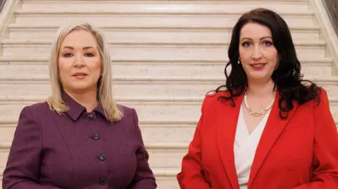 PA Michelle O'Neill and Emma-Little Pengelly stand side by side with the sandy stairs in Stormont behind them. O'Neill is wearing a dark purple button-up blazer, with shoulder length blonde hair and has makeup on. Emma-Little Pengelly is wearing a red blazer and a white top with a pearl and gold design necklace. She has long black hair and red lipstick on.