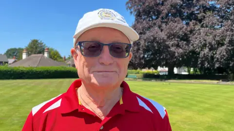 Richard looking into the camera and smiling slightly. He is wearing a red and white short-sleeved sports shirt and a white baseball cap. He is standing in front of a bright green, grassy bowling lawn, with a large tree visible over his right shoulder. The sky is bright blue and clear and it is very sunny.