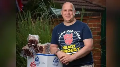 John McGall A bald man in a blue T shirt with an image on the front saying "I survived open heart surgery" leaning on a tall white box with three loaves of bread on top in front of a tall green plant and behind that a brick wall.