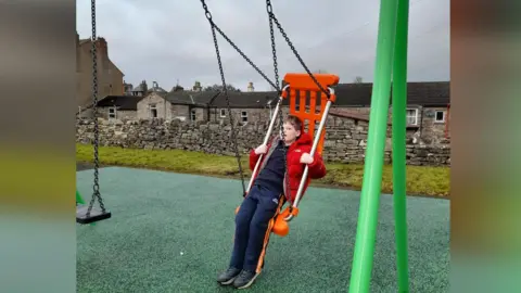 Alex on a large adapted swing in a playground. It has a green frame and orange seat. He is leaning on the seat and looking at the camera. He is wearing a navy blue tracksuit and red coat. He has short brown hair. Behind him is a drystone wall and some stone cottages.
