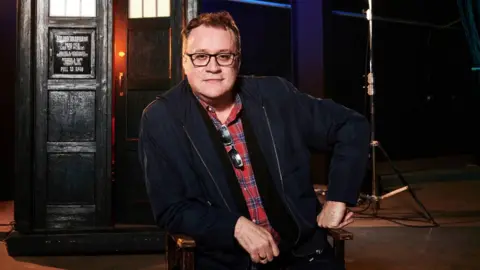 Russell T Davies is wearing a red and blue checked shirt with a black jacket. It's a professional photograph taken on the set of Dr Who. You can see the Tardis in the background. He has dark hair and wears black glasses. 