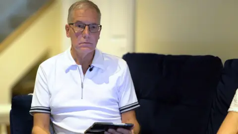 Marc Powell is pictured sitting on a blue sofa, operating a handheld computer tablet
