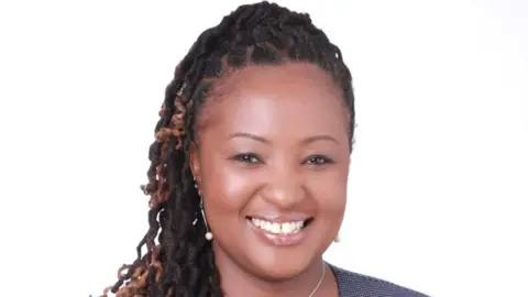 Metropolitan Police Ms Kilonzi is seen from the shoulders up against a white background. She has brown eyes and is smiling. Her hair is in a half up, half down do and has jewel drop earrings in. 