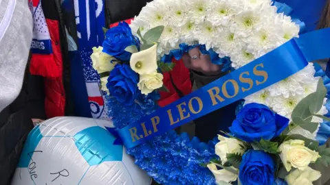 A wreath which reads RIP LEO ROSS. A ringed finger is holding it. Football scarves can be seen in the background. A balloon is also cropped. 