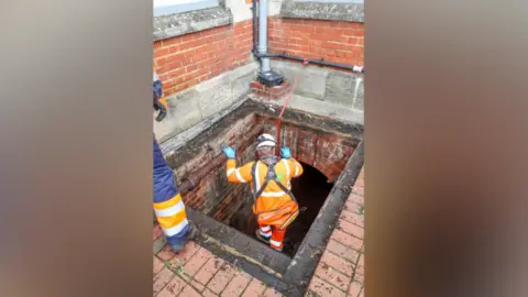 Spencer Mulholland/Salisbury Journal A large square bricked opening in the ground with a workman in a high-vis jacket being lowered in on a safety harness