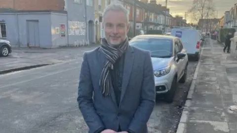 Councillor Paul Drake-Davis standing on a street in Hull wearing a suit and a scarf. Behind him are cars, houses and a tree in the distance.