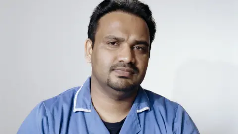 Daisy Moseley Portrait of Nidheesh Madhava. He has short dark hair and facial hair. He is wearing a blue care uniform with a collar. He is looking directly at the camera with a slight smile. 