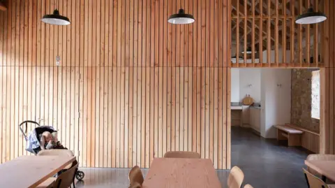 Emma Lewis Une pièce lambrissée avec trois tables à manger en bois entourées de chaises. Une ouverture dans le mur dessert un coin cuisine. Dans le coin de la pièce se trouve un bébé dans une poussette