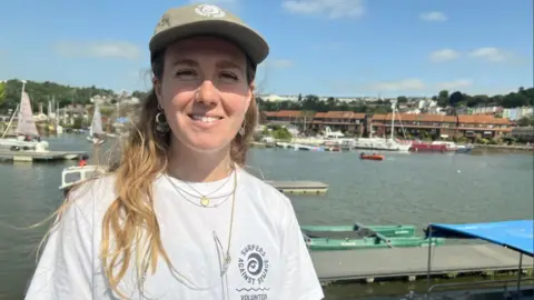 A woman with long fair hair and a green hat wearing a Surfers Against Sewage hat stands in front of a busy harbour. Sailing boats and power boats are visible in the background, alongside rows of houses on the far side of the water.