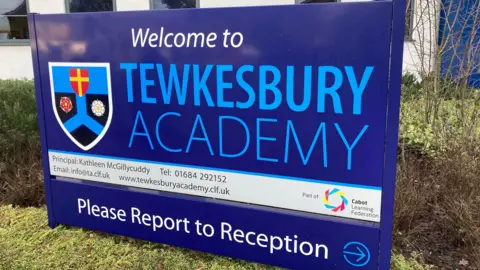 A large dark blue "welcome to Tewkesbury Academy" school sign on a grassy bank in front of a hedge and shrubs.