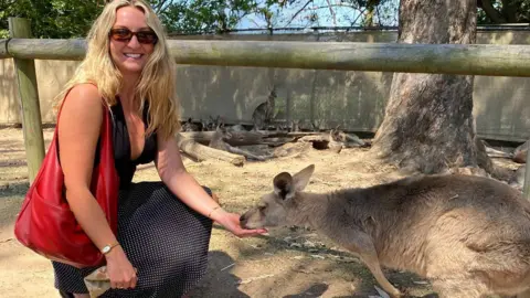 Chanel Buckland Fotografia de Chanel Buckland, 28 anos. Ela está ajoelhada diante de um canguru comendo na palma da sua mão. Ela tem cabelos loiros na altura dos braços, carrega uma bolsa vermelha e óculos escuros marrons. Ela usa uma saia preta de bolinhas e um top preto com decote redondo. Ao fundo você pode ver um grupo de cangurus. 