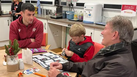 Rhianna Venables/BBC Goalkeeper Tom Smith sitting at a table with a man and a young boy. The men are chatting while smiling and the boy is putting stickers in a sticker book.
