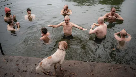 Ben Birchall/PA Wire A group of men standing in cold river water, with one man holding his arms outstretched, smiling at a dog stood on nearby steps.