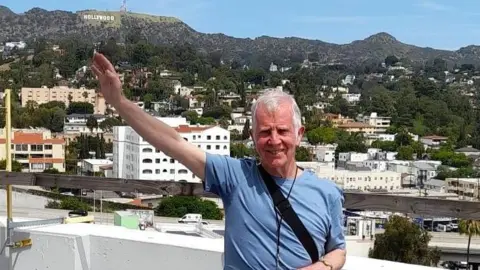 Jim, a mature man with white short hair and wearing a light blue T-shirt raises his arm towards the "Hollywood" sign as he checks out the roof on his tour of the Capitol Records building.