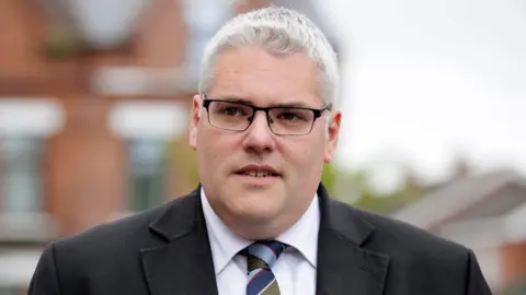 PA Media Gavin Robinson - a man with grey hair wearing black rimmed glasses and a dark suit jacket, white collared shirt and tie.