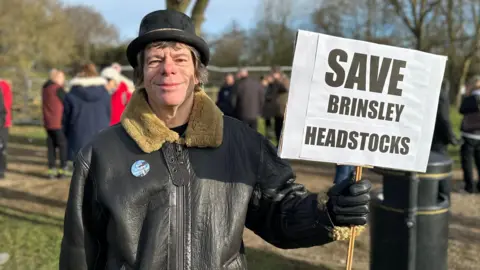 A grinning man in a leather jacket and a bowler hat holding a placard saying SAVE BRINSLEY HEADSTOCKS