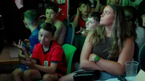 Jamie Niblock/BBC A young boy in a red England top shrugs his shoulders and raises his eyebrows. He is sitting watching the game with a group of other young boys and a woman.
