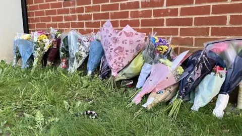 Flower tributes lying on grass propped up against a building.