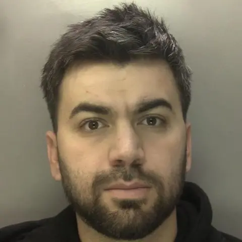 West Midlands Police A man with short dark hair and a dark beard, wearing a black hoodie, stands in front of a white wall, pictured from the shoulders up and looking at the camera.