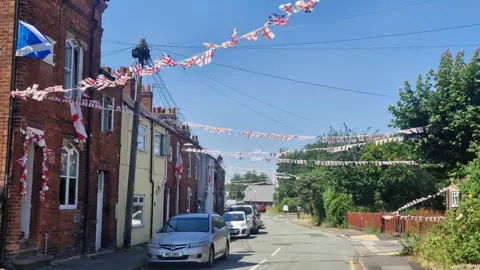 Mairead Smyth/BBC A view down a residential street lined with terraced houses. Streams of bunting with English and British flags cross the street while larger England flags hang from windows.