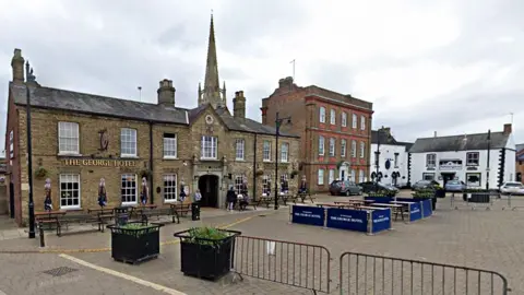 Google Whittlesey Market Place, showing the George Hotel and a church spire in the backdrop. There are people milling about and outdoor benches to eat on.