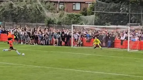 Derek Lloyd A Droylsden player, in a red shirt and black shorts, leans back as he takes the decisive penalty against a goalkeeper all in yellow, with fans packed behind the goal