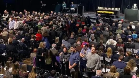 RTÉ A seated crowd in a large venue. Some members of the crowd are stood with their backs to the stage. One member of the crowd is being grabbed by a woman. 