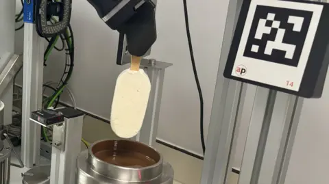 BBC A robot arm holds a Magnum ice cream above a pot of chocolate.