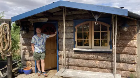 Ash Robertson stands in the doorway of his garden shed with one arm resting on the doorframe. He wears a Hawaiian shirt with green shorts. The shed has the look of a log cabin with a swing hanging in front of the window. 