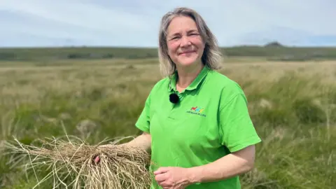 Joss Hibbs is smiling at the camera standing in opern moorland. She is wearing a bright green polo T-shirt. In her hand is a bundle of dead grass. 