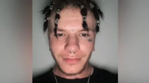 Supplied A man taking a photo of himself stood against a white background. He is wearing a black t-shirt, has a tattoo below his left eye, a nose ring and his hair tied in bunches