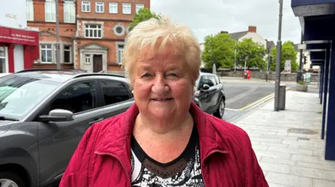 Marie Byrne, a woman with short blonde hair, smiles at the camera while standing on Hill Street in Newry. She is wearing a red quilted jacket over a black and white patterned top. Shop fronts and a tall red brick building - Newry Town Hall - are in the background. There are cars parked along the pavement.