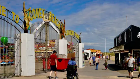 Entrance gates to amusement park with Coney Beach Pleasure Park in yellow letters on top of the gates and attractions visible behind