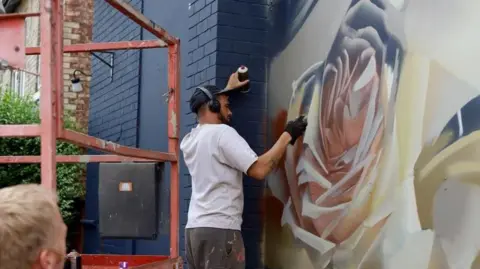 Art of Protest A male artist is painting a large mural on a wall. He is wearing a white shirt and black baseball cap with headphones on.