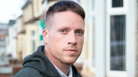 BBC A man with short brown hair and light stubble looks at the camera. Behind him are a row of terraced houses and a Welsh flag.