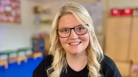 A woman with long blonde hair, smiling at the camera and wearing dark-rimmed glasses and and a black top.