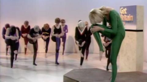 Diana Moran, wearing a green bodysuit, demonstrating an exercise move in front of a class of women.