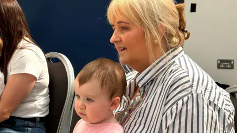 A woman with blonde hair in a bun wearing a white top with black stripes sits on a chair, holding a young child The child has short, dark hair and is wearing a pink top. The side of someone's arm is visible on the right of the pair.