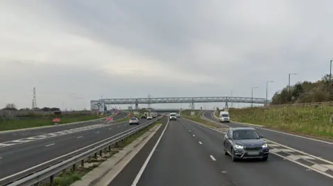 A street view of the A19 where the collision took place. It is a road with two lanes on each side with an additional lane on each side leading to a raised junction. A central reservation with metal barrier separates the carriageways. A silver footbridge goes across the road and the bridge at the top of the junction lies behind it. There are a number of vehicles on both sides but it is not busy.