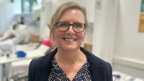 Close-up of Marjorie Dalton standing in the hospital ward in a navy blouse with white spots and a navy blazer. She is smiling, has blonde hair tied back and is wearing glasses.