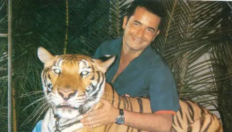 Acun Medya A person wearing a dark shirt crouches beside a tiger while posing for a photograph in front of palm leaves.