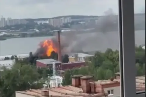 A still from a video taken from a window, showing an explosion over a factory-type building near a river